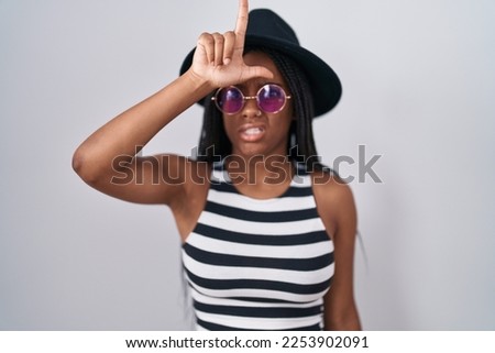 Young african american with braids wearing hat and sunglasses making fun of people with fingers on forehead doing loser gesture mocking and insulting. 