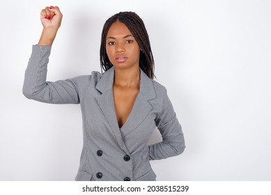 Young African American Business Woman With Braids Over White Wall Feeling Serious, Strong And Rebellious, Raising Fist Up, Protesting Or Fighting For Revolution.