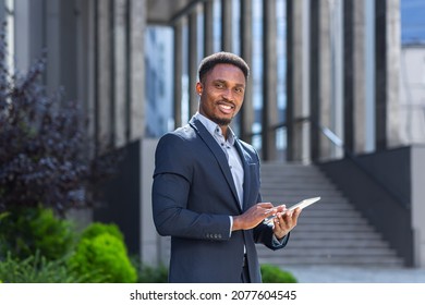 Young african american businessman in formal business suit standing working with tablet in hands on background modern office building outside. Man using smartphone or mobile phone outdoors city street - Shutterstock ID 2077604545