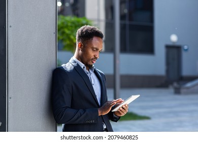 Young African American Businessman In Formal Business Suit Standing Working With Tablet In Hands On Background Modern Office Building Outside. Man Using Smartphone Or Mobile Phone Outdoors City Street