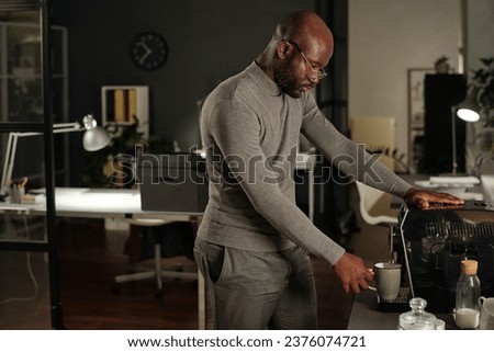 Young African American businessman in grey turtleneck making coffee in machine while standing in front of camera in spacious office at night