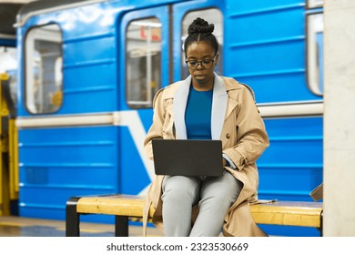 Young African American businesswoman with laptop sitting on bench against blue subway train and organizing work or using video chat