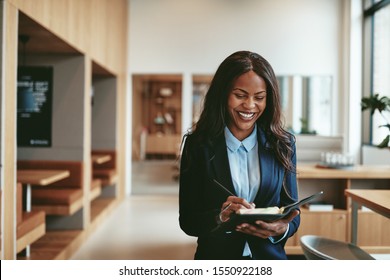 Young African American businesswoman laughing and writing notes in her day planner while walking in a bright modern office