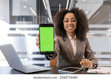 Young African American businesswoman in professional attire presenting smartphone with green screen in office. Confident woman smiling and demonstrating technology with laptop and documents on desk - Powered by Shutterstock