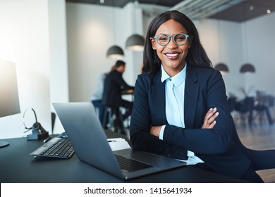 Young African American businesswoman smiling confidently while sitting with her arms crossed at her office desk with colleagues in the background - Powered by Shutterstock