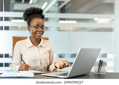 Young African American businesswoman working on laptop at workplace inside office, smiling confidently. Professional woman engaged in business tasks and online communication. - Powered by Shutterstock