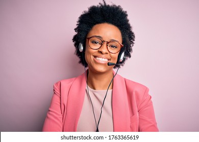 Young African American call center operator woman with curly hair using headset happy face smiling with crossed arms looking at the camera. Positive person.