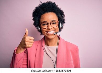 Young African American call center operator woman with curly hair using headset doing happy thumbs up gesture with hand. Approving expression looking at the camera showing success.