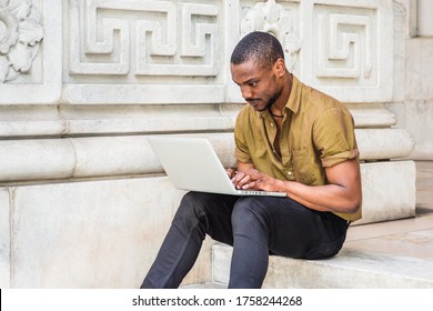 Young African American College Student Studying In New York City, Wearing Green Short Sleeve Shirt, Black Pants, Sitting On Stairs Outside Office Building On Campus, Working On Laptop Computer.