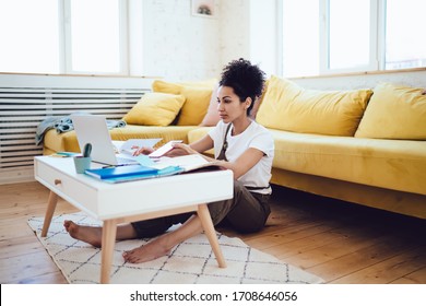 Young African American Concentrated Female Entrepreneur Working Remotely At Home Using Laptop And Sitting At Small Table On Floor Back Against Yellow Sofa In Living Room