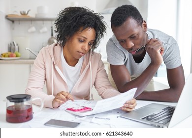 Young African American Couple Doing Paperwork Together, Planning Family Budget, Calculating Domestic Expenses, Sitting At Kitchen Table With Laptop And Calculator At Home. Financial Problems Concept