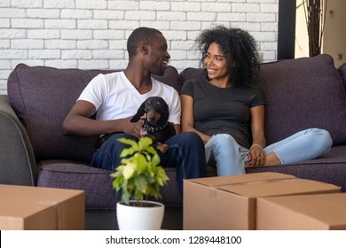 Young african american couple happy to move into new home with pet and boxes, black family tenants celebrate relocation sitting on couch with dog, homeowners renters having fun unpacking in own house