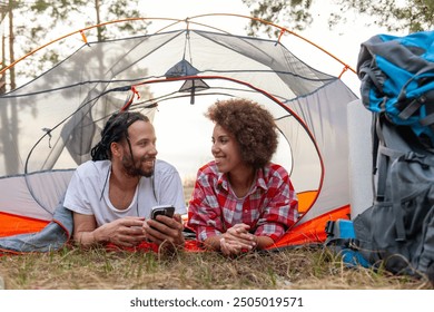 young african american couple on hike sitting and using smartphone in tent near lake in nature, man and woman tourists looking at mobile phone online on picnic trip and chatting - Powered by Shutterstock