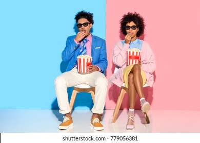 young african american couple sitting on chairs and eating popcorn against pink and blue wall