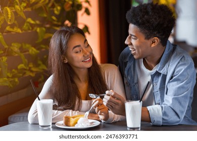 A young African American couple smiles warmly at each other while sharing a dessert and milkshakes at a charming outdoor cafe in the evening. - Powered by Shutterstock