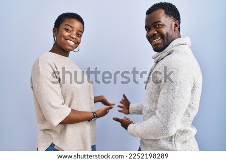 Young african american couple standing over blue background together inviting to enter smiling natural with open hand 