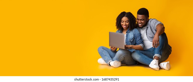 Young african american couple using laptop while sitting on floor over yellow background in studio, panorama with copy space