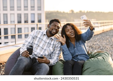 Young African American couple woman and man sitting on beanbag on rooftop of office during lunch and waving hands while taking selfie or having video call with mobile phone. - Powered by Shutterstock