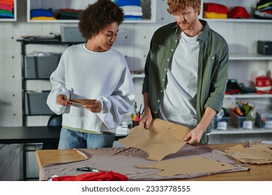 Young african american craftswoman holding sewing pattern and standing near cloth on table and redhead colleague in print studio, collaborative business owners working together - Powered by Shutterstock