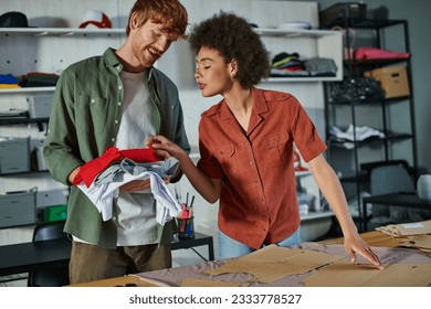 Young african american craftswoman looking at clothes in hands of cheerful colleague and working with fabric and sewing patterns in print studio, ambitious young entrepreneurs concept - Powered by Shutterstock