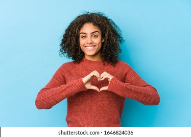 Young african american curly hair woman smiling and showing a heart shape with hands.