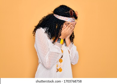 Young african american curly hippie woman wearing sunglasses and vintage accessories with sad expression covering face with hands while crying. Depression concept. - Shutterstock ID 1703768758