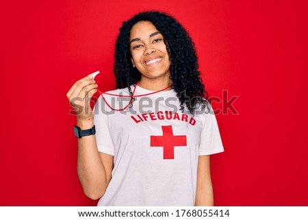 Young african american curly lifeguard woman wearing t-shirt with red cross using whistle with a happy face standing and smiling with a confident smile showing teeth