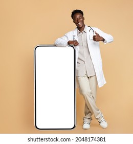 Young African American Doctor In Uniform Leaning At Big Smartphone With Blank Screen And Showing Thumb Up At Camera, Excited Black Male Medical Worker Demonstrating Free Copy Space, Mockup Image