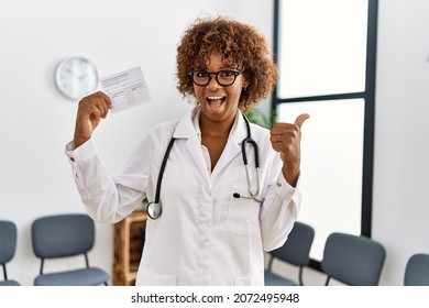 Young African American Doctor Woman Holding Covid Record Card Pointing Thumb Up To The Side Smiling Happy With Open Mouth 