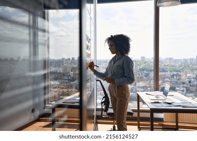 Young African american executive manager businesswoman writing strategy ideas on sticky notes on whiteboard doing business project presentation in contemporary corporate office with panoramic view.