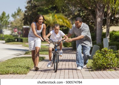 A Young African American Family With Boy Child Riding His Bicycle And His Happy Excited Parents Giving Encouragement Next To Him