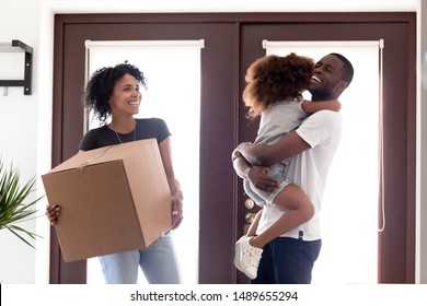 Young African American Family Stand At Doorway With Boxes In Hands Moving To New Home, Excited Small Girl Hug Smiling Dad Happy To Move Into Own House Or Apartment. Ownership Concept