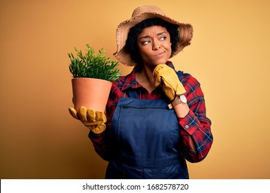 Young African American Farmer Woman With Curly Hair Wearing Apron Holding Pot With Plants Serious Face Thinking About Question, Very Confused Idea