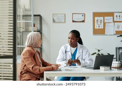 Young African American female clinician in lab coat talking to Muslim patient in hijab and casual apparel during medical consultation - Powered by Shutterstock