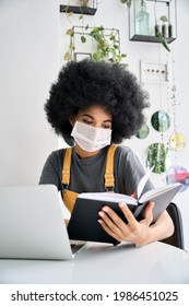 Young African American Female College Or University Student With Afro Hair Sitting In Cafe Indoor Remotely Studying Reading With Laptop Wearing Surgical Face Mask For Covid 19 Prevention.