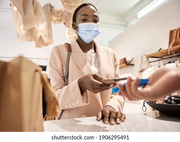 Young African American Female Customer Wearing A Protective Face Mask Making A Contactless Payment Using Her Smart Phone In A Clothing Store