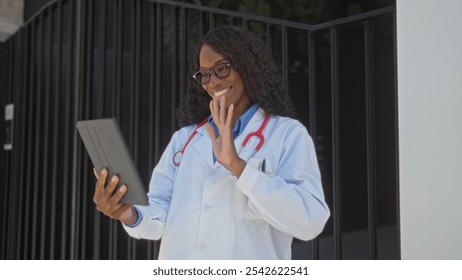 A young african american female doctor with curly hair is making a vide call outdoors in an urban street near a clinic, waving and smiling while holding a tablet. - Powered by Shutterstock