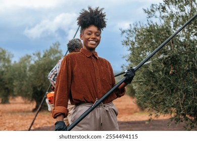 Young african american female farmer holding harvesting tool and smiling in an olive grove, with a coworker using a harvesting machine in background - Powered by Shutterstock