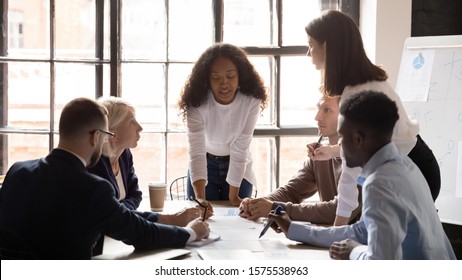 Young African American Female Professional Leaning Over Table, Explaining Marketing Data Analysis Paper Report To Concentrated Successful Mixed Race Coworkers At Brainstorming Meeting In Office.