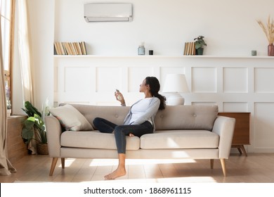 Young African American Female Renter Sit Rest On Sofa At Home Turn Air Conditioner Breathe Fresh Air. Millennial Biracial Woman Tenant Relax On Couch Switch AC Device With Remote. Technology Concept.