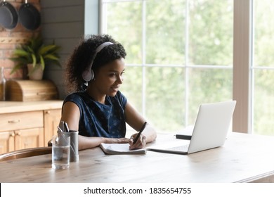 Young African American female student sit at table at home study distant on computer make notes. Millennial biracial woman in headphones watch webinar on laptop, work online or take web course.