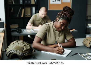 Young African American female student making notes in notepad after teacher while sitting by desk at lesson of military training