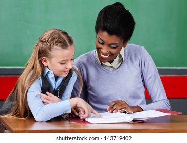 Young African American Female Teacher Assisting Schoolgirl At Desk In Classroom