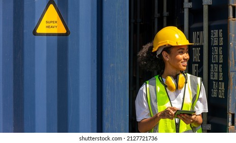 Young African American Female Trainee Check Cargo Containers And Stocks In A Logistic Warehouse. Female Worker In Hardhat And Safety Suit Stands Next To A Blue Container. Factory Driver With Earmuff