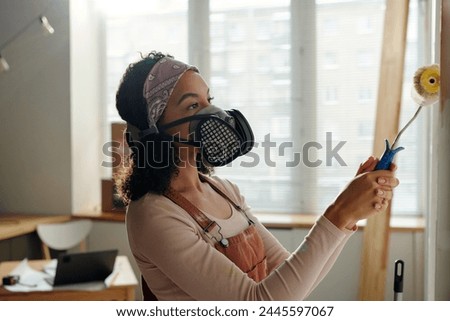 Young African American female worker in overalls and protective respirator holding by handle of paintroller while painting wall of cafe in white