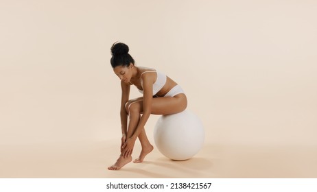 Young African American Fit Cute Brunette Woman With Bun Hairstyle In White Underwear Strokes Her Leg Sitting On Big White Ball On Beige Background | Leg Care Concept