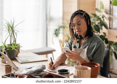 young african american florist in eyeglasses looking at smartphone and writing order in notebook in flower shop