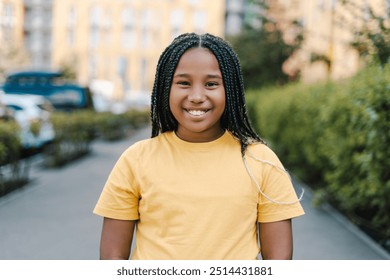 Young African American girl with braids is smiling while standing on a city street with greenery and a blurred apartment building in the background. Natural beauty concept - Powered by Shutterstock