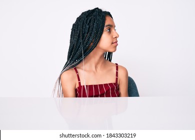 Young African American Girl Child With Braids Wearing Casual Clothes Sitting On The Table Looking To Side, Relax Profile Pose With Natural Face With Confident Smile. 