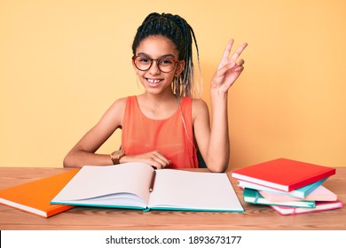 Young African American Girl Child With Braids Studying For School Exam Smiling With Happy Face Winking At The Camera Doing Victory Sign. Number Two. 